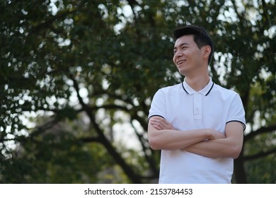 Asian Young Man In White Short Sleeve Polo Shirt, Cross His Arms, Smile And Look Away In Front Of Blur Green Tree