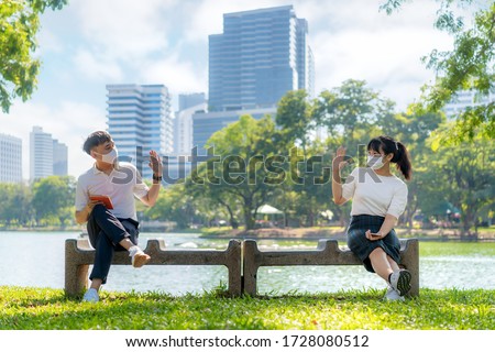 Asian young man and woman greet and say hello with they friend and wearing mask sitting distance of 6 feet distance protect from COVID-19 viruses for social distancing for infection risk in park.
