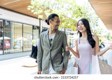 Asian Young Man And Woman Shopping Goods Outdoors In Department Store. Attractive Couple Hold Shopping Bags Then Walking Together With Happiness, Enjoy Purchasing In Shopping Mall Marketplace Center.