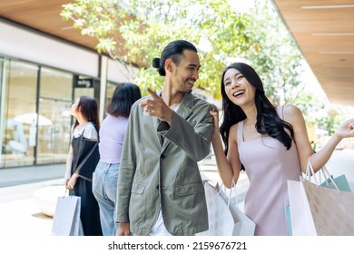 Asian Young Man And Woman Shopping Goods Outdoors In Department Store. Attractive Couple Hold Shopping Bags Then Walking Together With Happiness, Enjoy Purchasing In Shopping Mall Marketplace Center.