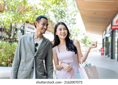 Asian Young Man And Woman Shopping Goods Outdoors In Department Store. Attractive Couple Hold Shopping Bags Then Walking Together With Happiness, Enjoy Purchasing In Shopping Mall Marketplace Center.