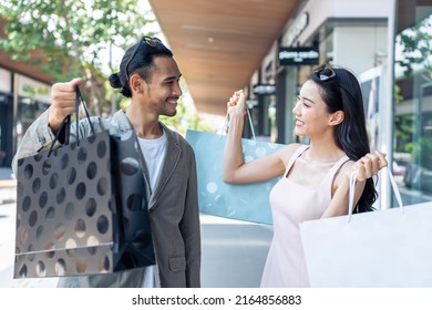 Asian Young Man And Woman Shopping Goods Outdoors In Department Store. Attractive Couple Hold Shopping Bags Then Walking Together With Happiness, Enjoy Purchasing In Shopping Mall Marketplace Center.