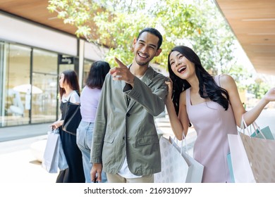 Asian Young Man And Woman Shopping Goods Outdoors In Department Store. Attractive Couple Hold Shopping Bags Then Walking Together With Happiness, Enjoy Purchasing In Shopping Mall Marketplace Center.