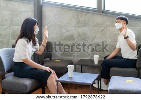 Asian young man and woman sitting one person per one table and greet and say hello for distance of 6 feet distance protect from COVID-19 viruses for social distancing for infection risk at coffee cafe