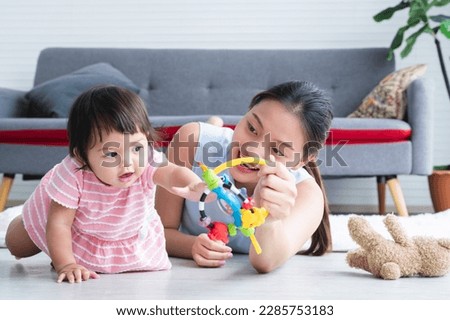 Asian young mother and Caucasian 7 months baby newborn girl playing with development toy and bear doll. Single mom lying on floor smiling playing with little daughter, crawling in living room at home