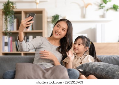 Asian Young Mother And Her Daughter Have Video Call Conference With Family Having Fun Together.Happiness Mom And Little Girl Looking At Mobile Phone And Waving Hand With Video Chat To Grandmother 