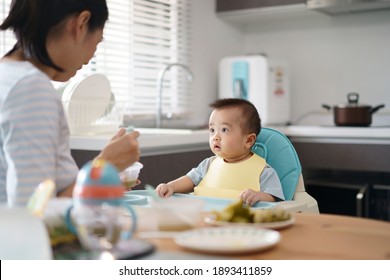 Asian young mother provide food and fruit puree for lunch to her baby seat on high chair in kitchen at home. The baby try to weaning and looking at his mom. A wife  take care her son.