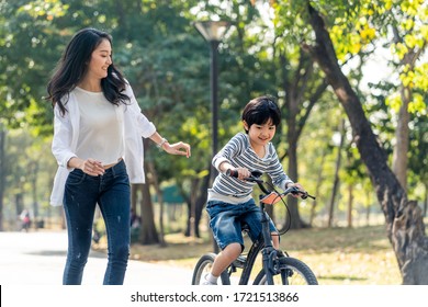 Asian young mother taking care kid while practicing to ride a bicycle by running follow son in public park. Child smiling to mom with happy face enjoy playing activity together. Boy feels fun to bike.