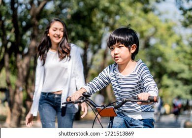 Asian Young Mother Taking Care Kid While Practicing To Ride A Bicycle By Running Follow Son In Public Park. Child Smiling To Mom With Happy Face Enjoy Playing Activity Together. Boy Feels Fun To Bike.