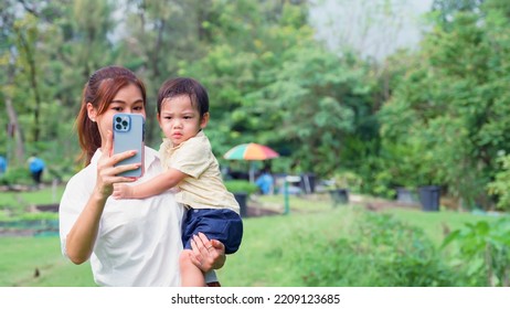 Asian Young Mother Taking A Selfie With A Cute Little Son Her In The Park While Her Son Was Irritated And Overbearing.