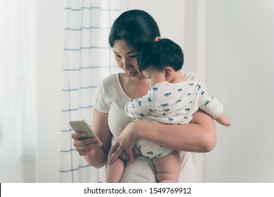 Asian Young Mother Working And Holding Baby While Talking On Phone In Home Office