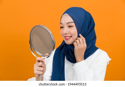 Asian young Muslim woman wearing hijab headscarf smiling and looking at mirror on orange colour background in studio - Powered by Shutterstock