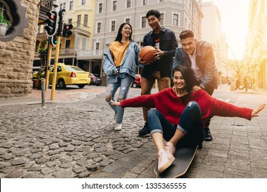 Asian Young People In The City With Skateboard And Basketball. Woman Being Pushed By Man  On Skateboard Outdoors On Street, With Their Friends Walking By In The City Street.