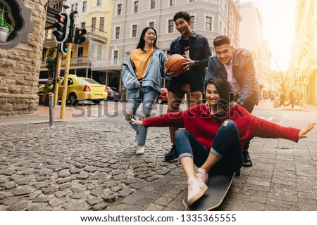 Asian young people in the city with skateboard and basketball. Woman being pushed by man  on skateboard outdoors on street, with their friends walking by in the city street.