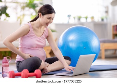 Asian Young Pregnant Woman Using Laptop To Have Online Exercise Class With Pilates Ball And Dumbbells In The Living Room