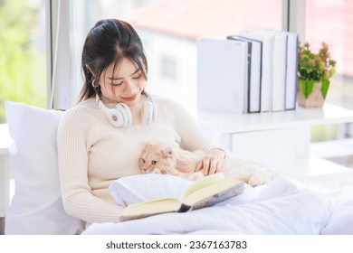 Asian young pretty cheerful female teenage girl in cute dress sitting on white clean sheet blanket bed smiling playing hugging holding cute domestic short hair pet cat companion friend in bedroom. - Shutterstock ID 2367163783