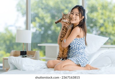 Asian young pretty cheerful female teenager girl in  dress sitting on white clean sheet blanket bed smiling playing hugging holding cute domestic short hair pet cat companion friend in bedroom. - Shutterstock ID 2367163815