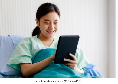 Asian Young Pretty Female Patient Smiling With Happiness And Touching Screen Of Tablet For Online Chat Or Communicate While Lying On Bed In Hospital. Technology And Medical Concept