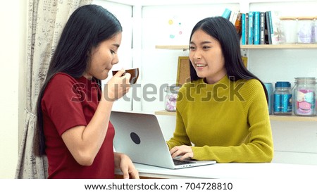 Asian young pretty woman using laptop computer to working and her friend come in with hot coffee and standing nearby, talking with joyful, teenager lifestyle concept