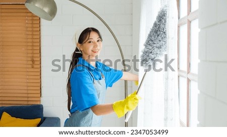 Asian young professional cleaning service woman worker working in the house. The girl cleans the curtain and window with a grey feather duster. Professional cleaner and chore service concept.