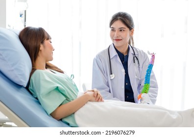 Asian Young Professional Female Doctor In White Lab Coat With Stethoscope Sitting Holding Human Spine Skeleton Model Showing Explaining Supporting Comforting Patient Laying Down On Bed In Ward Room.