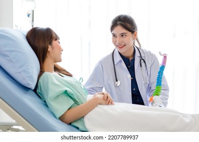 Asian Young Professional Female Doctor In White Lab Coat With Stethoscope Sitting Holding Human Spine Skeleton Model Showing Explaining Supporting Comforting Patient Laying Down On Bed In Ward Room.