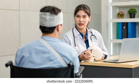 Asian Young Professional Female Practitioner Doctor In White Lab Coat With Stethoscope Holding Skull Model Showing Explaining To Unrecognizable Male Patient Sitting On Wheelchair After Brain Surgery.