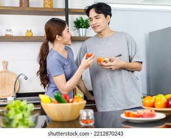 Asian Young Romantic Couple Beautiful Wife Standing Smiling Feed Sliced Mixed Fruits From Glass Bowl To Handsome Husband Behind Kitchen Counter Full Of Fresh Raw Organic Vegetables