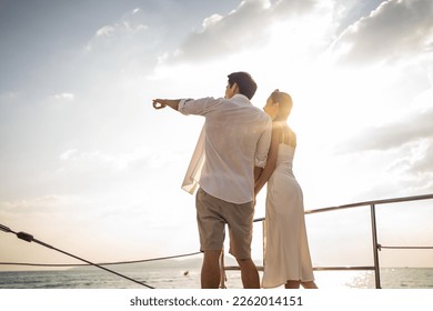 Asian young romantic couple looking at beautiful view during yachting. Attractive man and woman hanging out celebrating anniversary honeymoon trip while catamaran boat sailing during summer sunset.