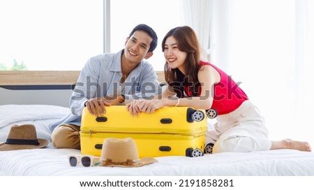Asian young romantic lover couple boyfriend and girlfriend in casual outfit sitting smiling helping packing luggage together on bed while male using tablet computer booking vacation trip hotel online.