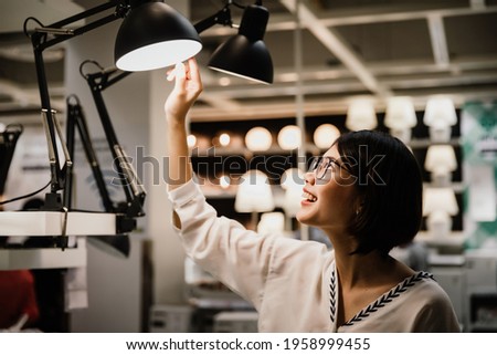 Asian young short hair beautiful woman wearing glasses choosing new lamp to buy at furnishings lighting store