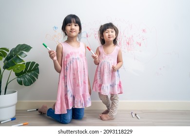Asian young sibling kid girl enjoy paint on white wall in living room. Little adorable children having fun drawing and coloring art picture with hapiness enjoy creativity at home and looking at camera