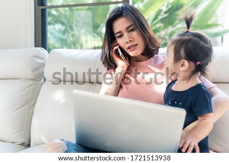 Asian young single mother talking on mobile phone and using computer for working from home while take care little baby girl together at home. Woman busy with urgent work. Due to Covid19 virus pandemic