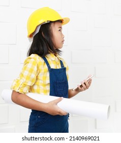Asian young smart clever little preschooler girl future dream job career as engineering and architecture wears yellow safety hardhat helmet standing on call with smartphone holding blueprint in hand.