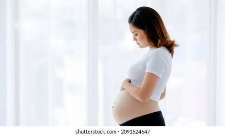 Asian Young Stressed Depressed Upset Sick Unhappy Female Pregnancy Mother Standing Alone Thinking Worrying About Unborn Baby Holding Touching Naked Belly Tummy Stomach In Front Curtain Background.