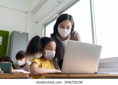 Asian Young Teacher With African American Girl In Protective Face Masks Studying On Laptop In Classroom. Woman Teaching Diversity Students In International School. Education And Learning On Technology