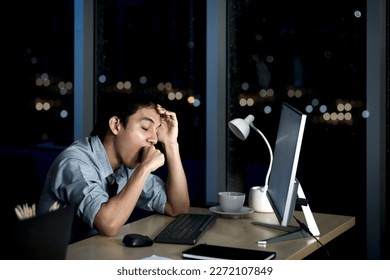 Asian young tired staff officer yawning during using desktop computer, feeling sleepy after having overwork project overnight, exhausted businessman working hard overtime at night in office workplace. - Shutterstock ID 2272107849
