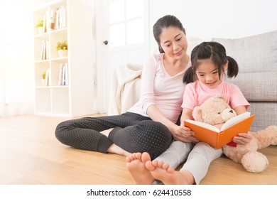 asian young woman accompany children reading comic book in the afternoon of the holiday at home in the living room and lovely girl holding her teddy bear sitting on the floor to earnestly study.