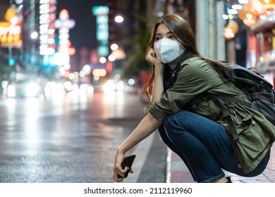 Asian Young Woman Backpacker Wear Mask, Wait For Bus In City At Night. Beautiful Tourist Traveler Girl Travel Alone On Street With Colorful Light, Enjoy Spend Time On Holiday Vacation During Pandemic.