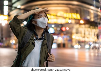 Asian Young Woman Backpacker Wear Mask, Wait For Bus In City At Night. Beautiful Tourist Traveler Girl Travel Alone On Street With Colorful Light, Enjoy Spend Time On Holiday Vacation During Pandemic.