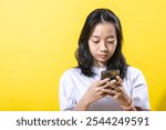 An Asian young woman with black hair wearing a white t-shirt and brown pants, holding a smartphone with both hands. She is focused on the phone, against a vibrant yellow background