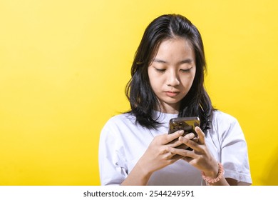An Asian young woman with black hair wearing a white t-shirt and brown pants, holding a smartphone with both hands. She is focused on the phone, against a vibrant yellow background