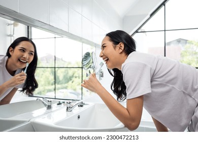 Asian young woman brush teeth and look at mirror in bathroom at home. Attractive beautiful female brushing teeth with toothbrush and toothpaste enjoying morning oral anticavity hygienic routine alone. - Powered by Shutterstock