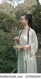 An Asian Young Woman Dressed Up In Vintage Style Holding A Bamboo Basket In A Traditional China Garden. 