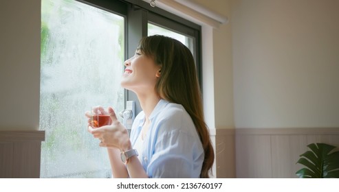 Asian Young Woman Is Drinking Tea Or Coffee While She Looking Through Window In A Rainy Day At Home