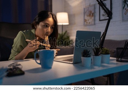 Asian young woman eating instant noodles while watching video movie on tablet and reading news, overtime night, late time at home