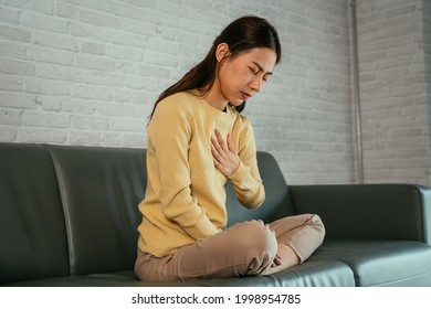 Asian Young Woman Feeling Discomfort As Suffering From Heartburn Holding Chest With Closed Eyes And Sitting With Folded Legs On Couch At Home