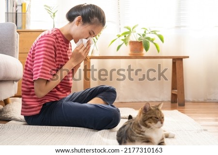 Asian young woman, girl hand in sneezing from fur allergy while playing with her lovely cat, pet on carpet in living room at home, apartment. Health care rhinitis diseases allergic to animal hair.