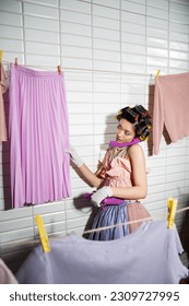 asian young woman with hair curlers standing in pink ruffled top, pearl necklace and white gloves, talking on purple retro phone and looking at wet skirt near clean laundry handing near white tiles