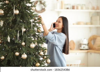 Asian Young Woman Has A Conversation On Mobile Phone While Decorating Christmas Tree At Home